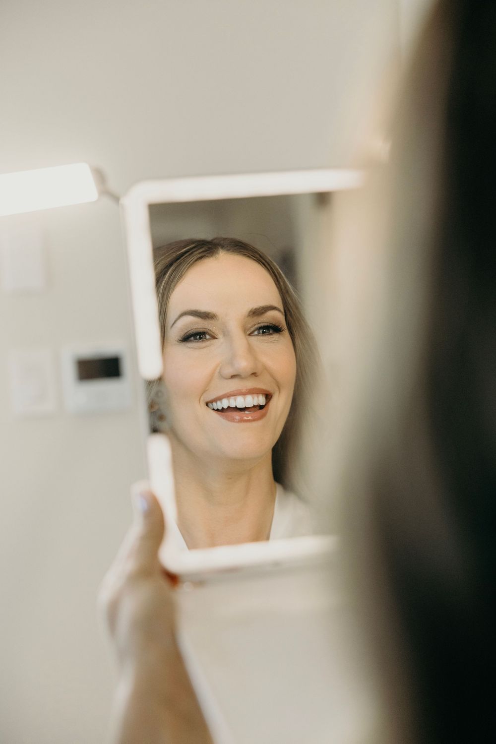 Close up reflection of a smiling person in a mirror being held up during wedding preparations.