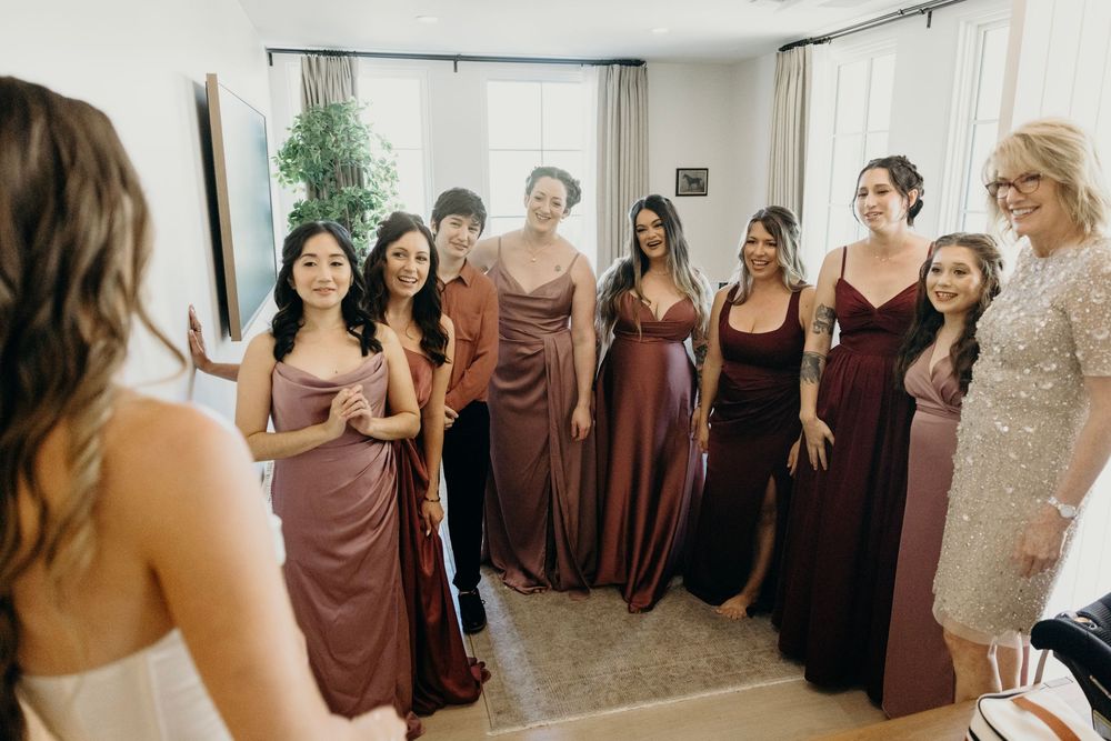 Wedding party members in burgundy and black dresses standing together in a sunlit room.