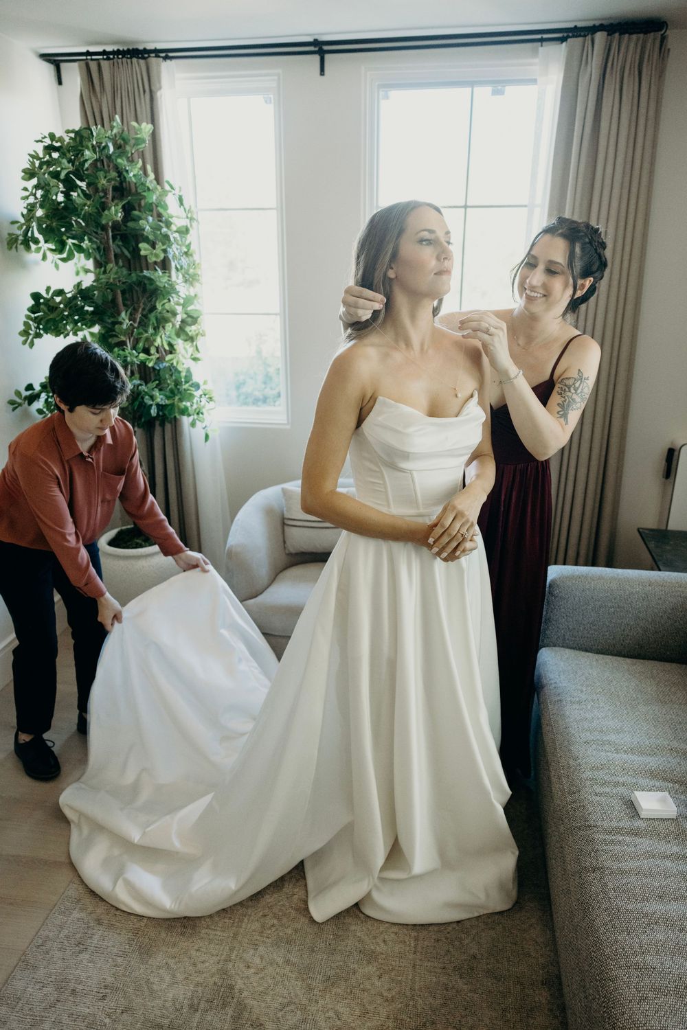 Bride in white wedding dress getting help with final adjustments in a bright room.