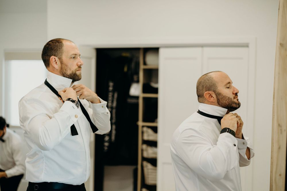 Men in white shirts adjusting black bow ties before wedding ceremony.
