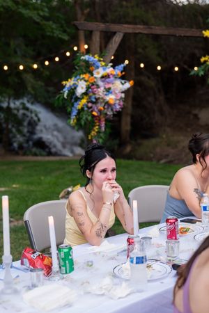 People seated at an outdoor dining table with string lights and beverages during a casual gathering.