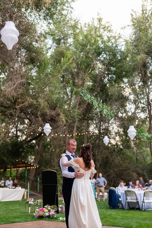 Romantic outdoor garden wedding reception with string lights and white lanterns hanging among trees at dusk.