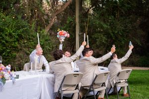 Wedding party in light gray suits raises champagne glasses in celebration at an outdoor reception table.
