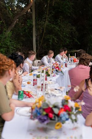 Guests seated at a long outdoor wedding reception table with colorful floral centerpieces and refreshments.