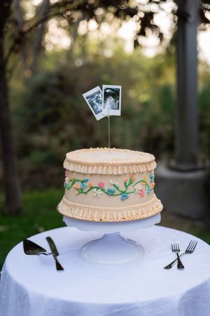 A vintage-style wedding cake with floral embroidery design sits on a white tablecloth with decorative photos on top.