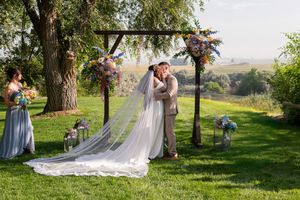 Wedding party gathers under a tree in a scenic outdoor setting during a romantic countryside ceremony.