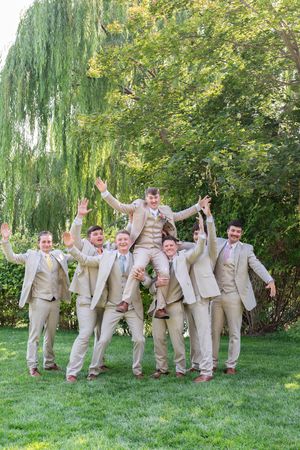 Wedding party in tan suits strike fun poses on green lawn with willow trees in background.