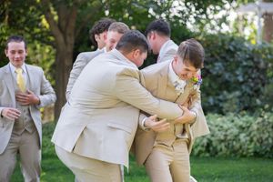 Groomsmen in tan suits perform synchronized dance moves at an outdoor wedding reception.