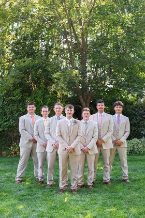 A group of groomsmen in light beige suits pose together on a grassy lawn with green trees in the background.