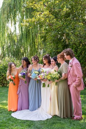 Bridesmaids in pastel dresses of orange, purple, blue, cream and pink stand in line holding colorful bouquets outdoors.