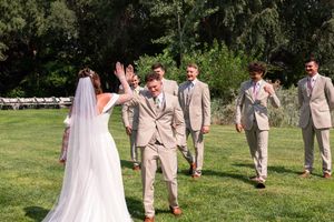 Wedding party members in beige suits and a flowing white dress pose playfully in a sunny outdoor garden setting.