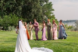 Wedding party gathers on a lush green lawn overlooking water, with bridesmaids in pastel dresses watching the bride.