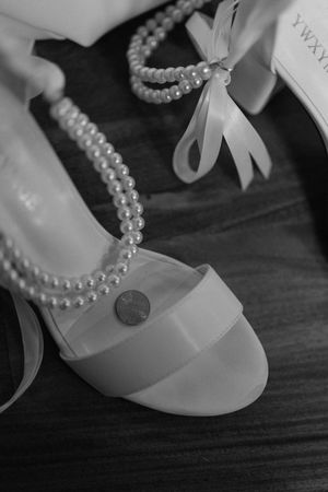 Black and white photo of elegant white bridal shoes with pearl necklace and delicate bow details on wooden surface.