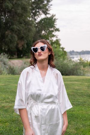 A person in a white satin top and sunglasses poses playfully outdoors on a grassy hillside.
