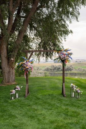 Rustic wooden wedding arch decorated with colorful wildflowers stands on green lawn with lanterns beneath willow trees.