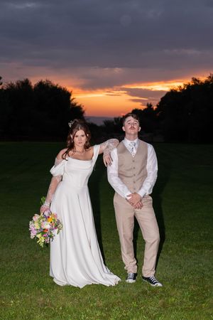 A couple poses playfully in a grassy field at sunset wearing wedding attire and casual shoes.