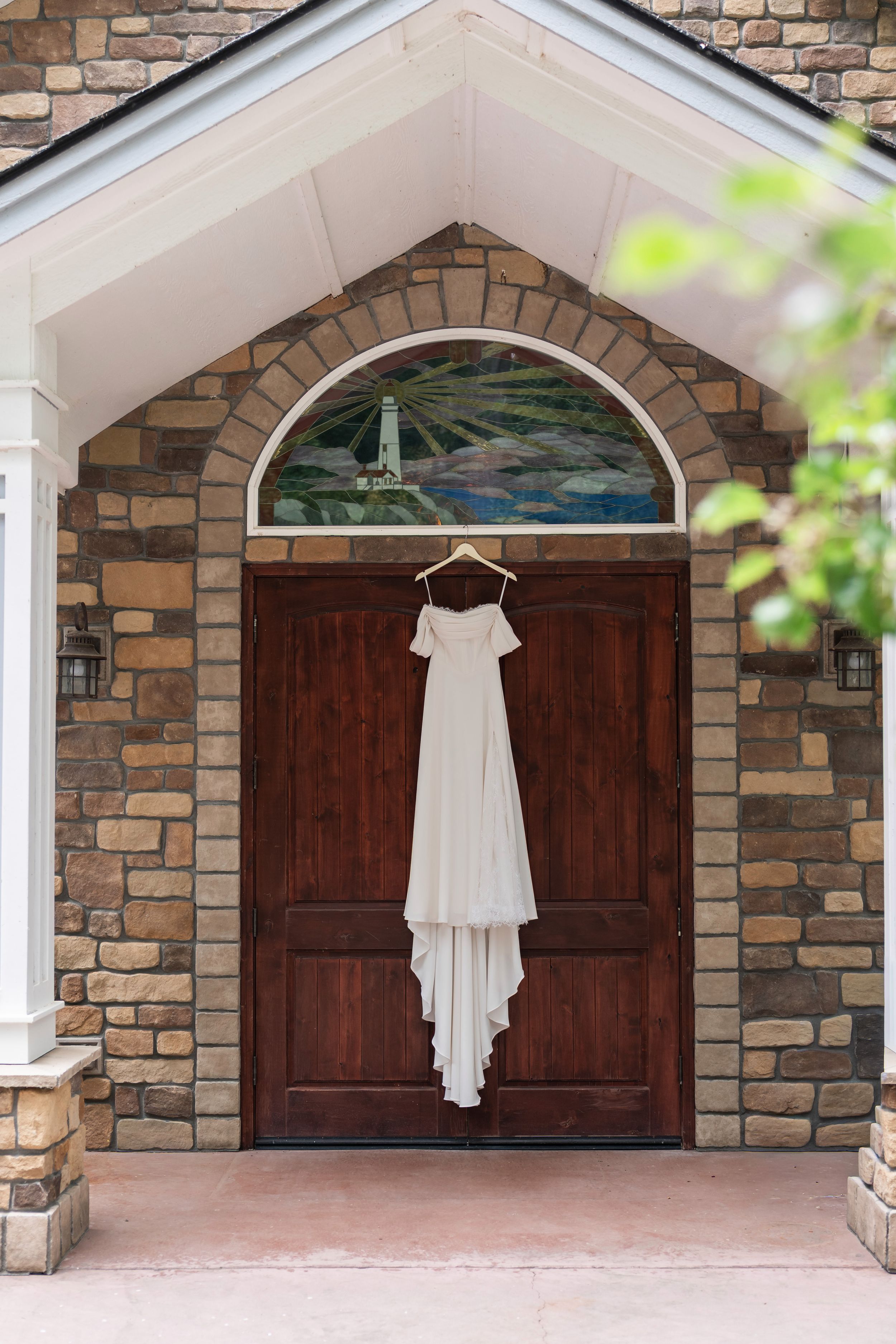 White wedding dress hanging elegantly against dark wooden doors with stone archway and brick exterior.