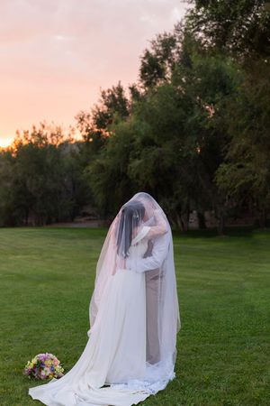 A bride in a flowing white veil and dress stands in a green field at sunset with trees in the background.