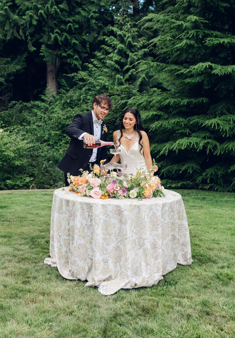 A couple cuts their wedding cake at an outdoor reception with a beautifully decorated table against evergreen trees.