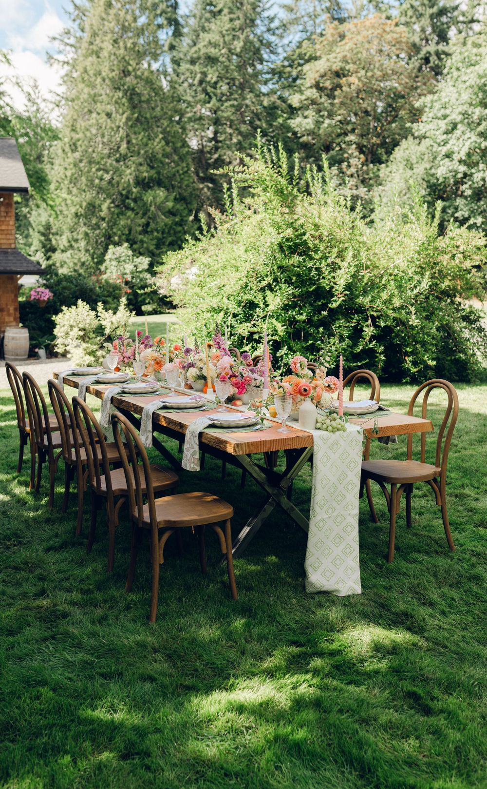 Long wooden dining tables decorated with floral arrangements set up outdoors on a grassy lawn under trees.