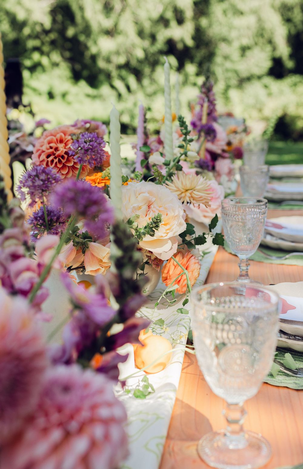 Elegant table setting with crystal glasses and soft peach and purple floral arrangements at an outdoor garden party.