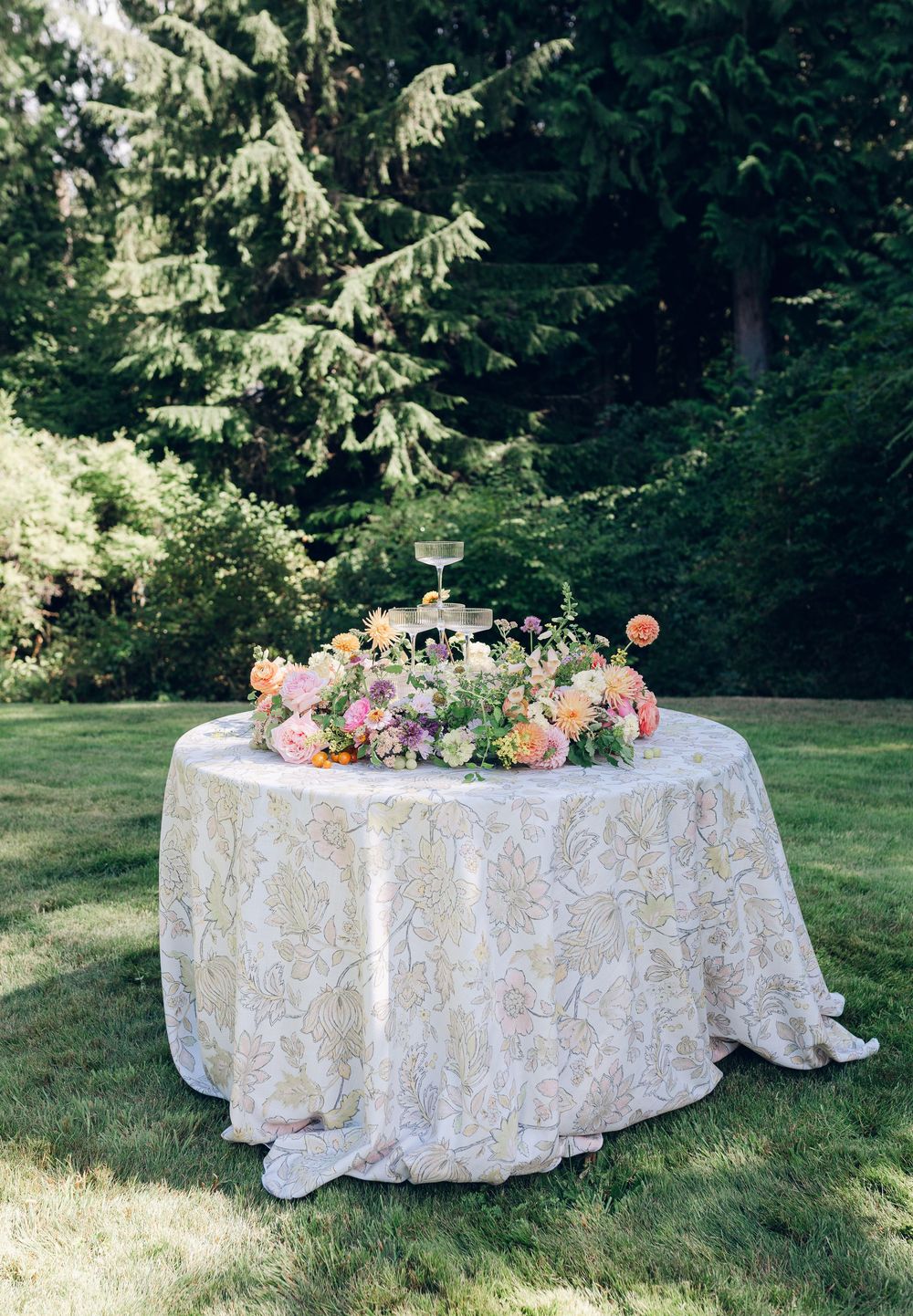 Elegant garden table setup with lace tablecloth and pink floral centerpiece arrangements on a sunny lawn with evergreens.