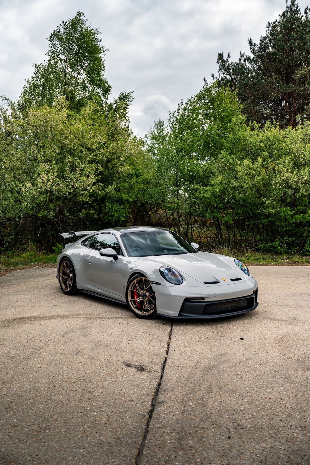 A sleek silver Porsche 911 GT3 sports car parked on a gravel surface with lush green trees in the background.