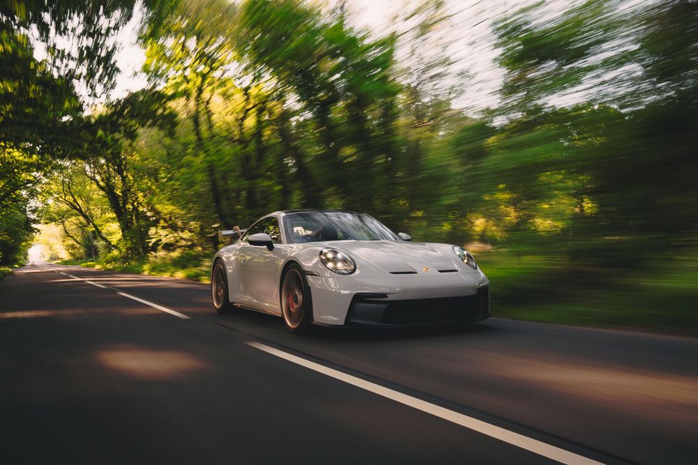 Motion-blurred shot of a silver Porsche GT3 driving on a winding forest road at sunset.