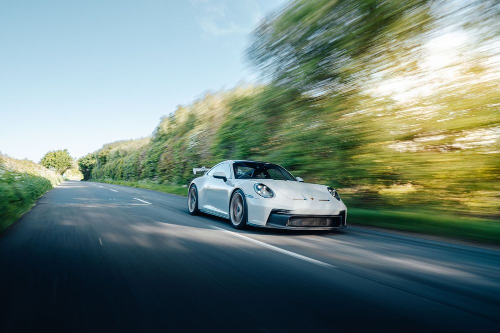 White sports car driving on a winding road with motion blur effect and green trees in the background.