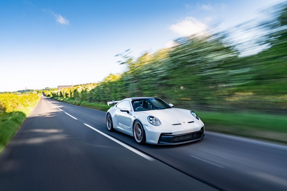 White Porsche sports car driving on a winding road with motion blur and scenic landscape in the background.