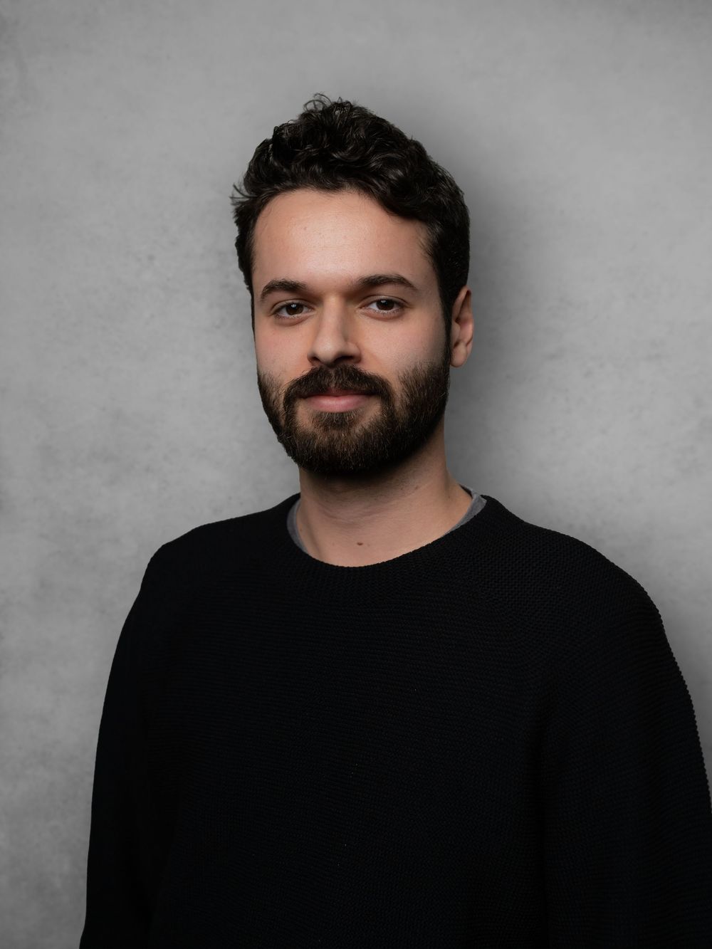 Professional headshot of person in black shirt against gray background with dark beard and styled hair.