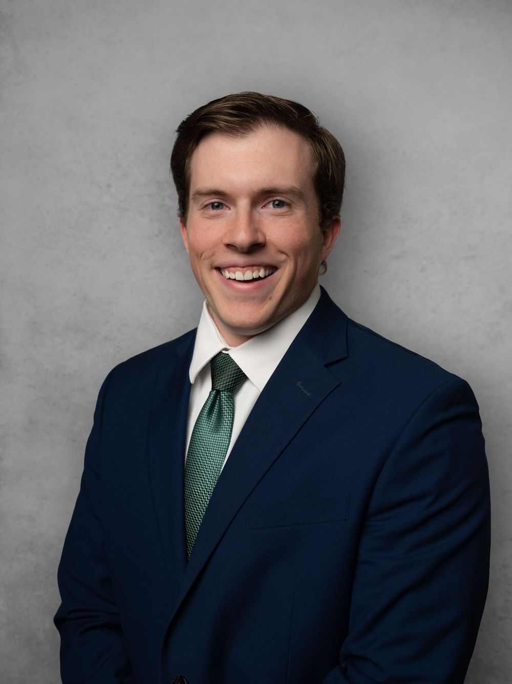 Professional business portrait of smiling person in navy suit with green tie against gray background.