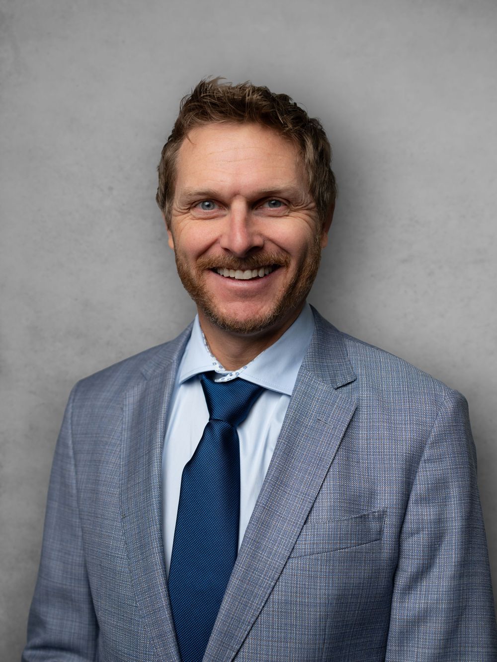 Professional headshots of someone in a gray suit and blue tie against a neutral background.