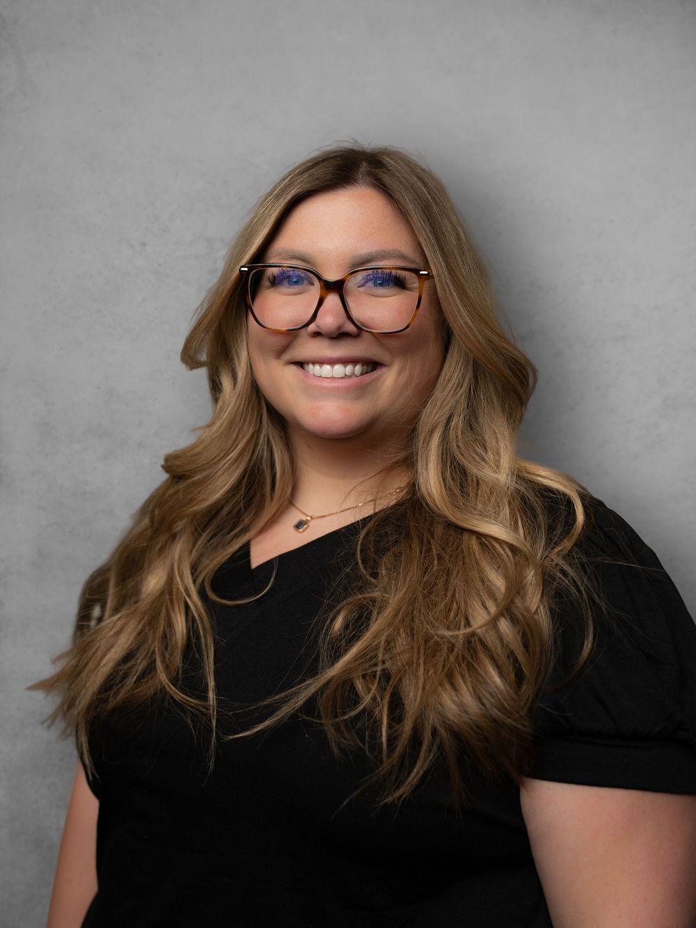 Professional portrait of someone with wavy blonde hair, glasses and a black top against a gray background.