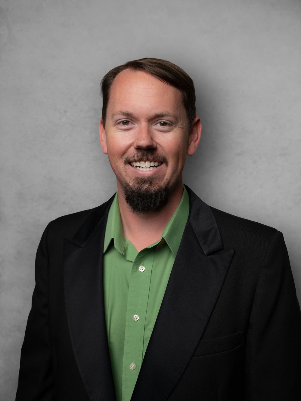 Business portrait of person wearing green polo shirt and black blazer against neutral backdrop.