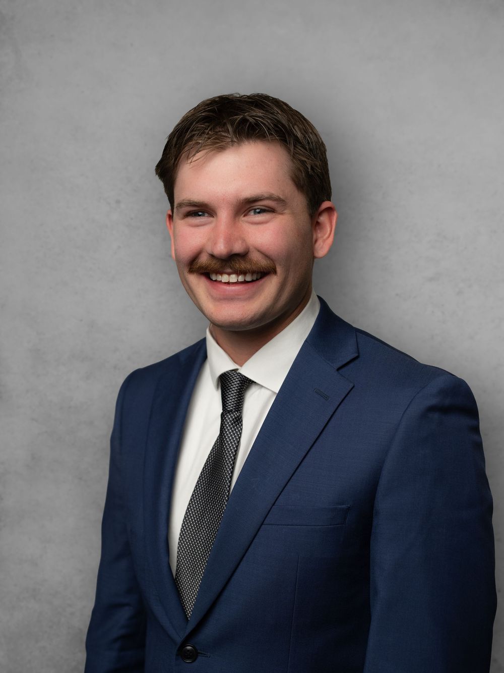 Professional headshot of person in navy suit and dark tie against gray background.