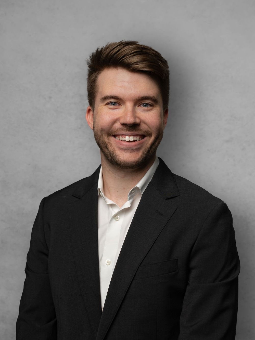 Professional headshot series of a person in a black suit and white shirt against a gray background showing different expressions.