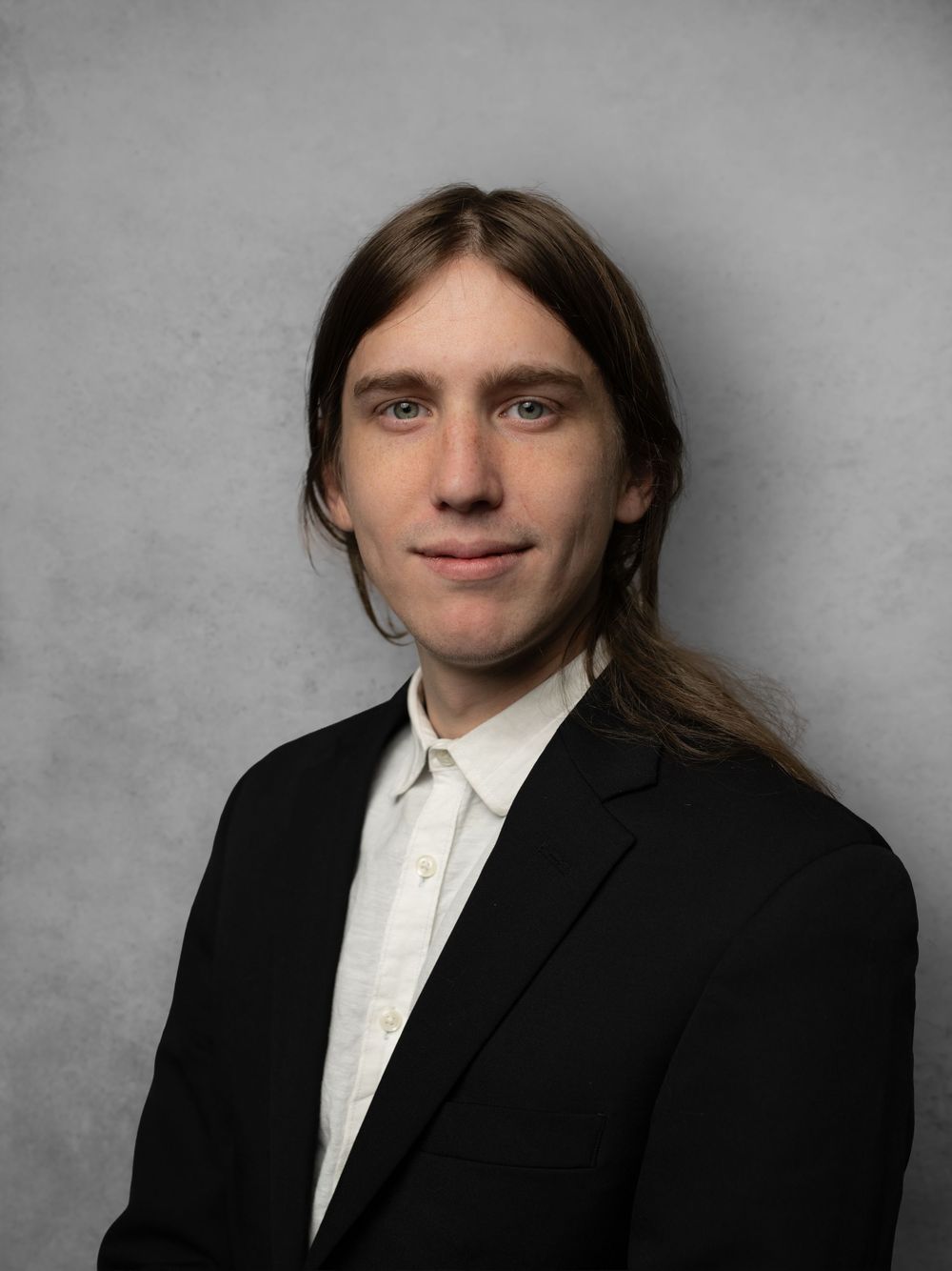Portrait of person with long hair wearing black suit and white shirt against gray backdrop.