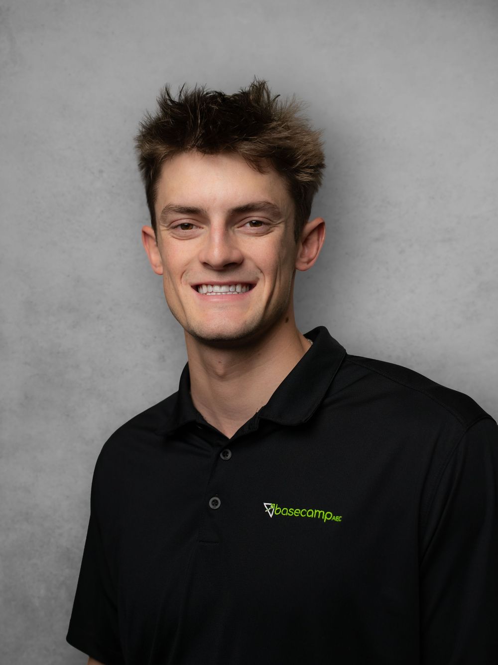 Person in black polo with green logo smiling against gray background.