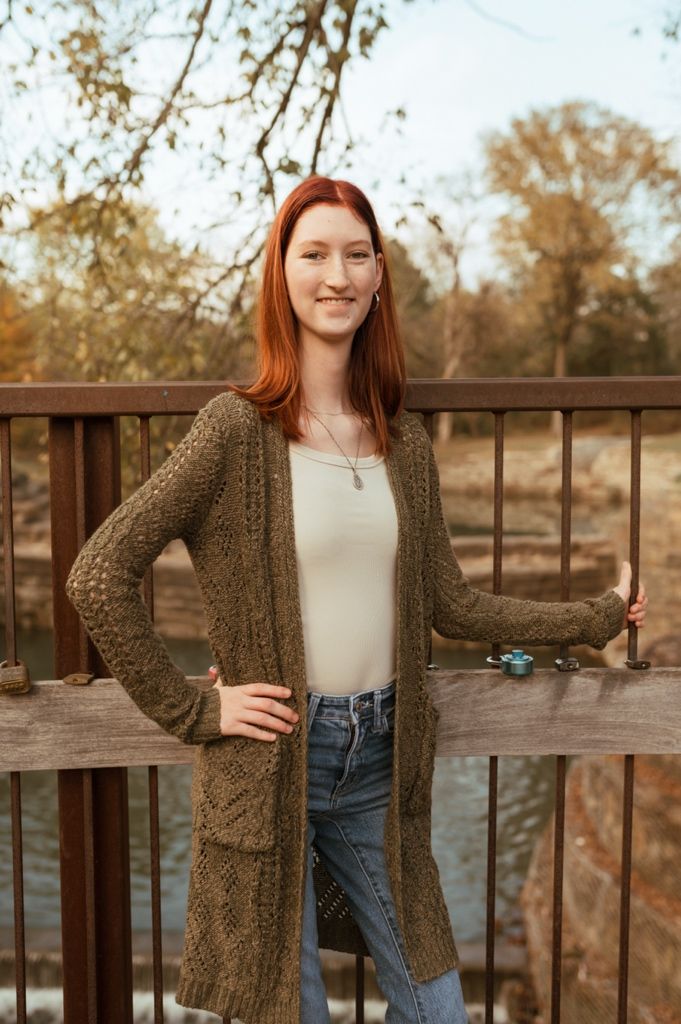 A person in a brown cardigan poses casually against a wooden bridge railing.