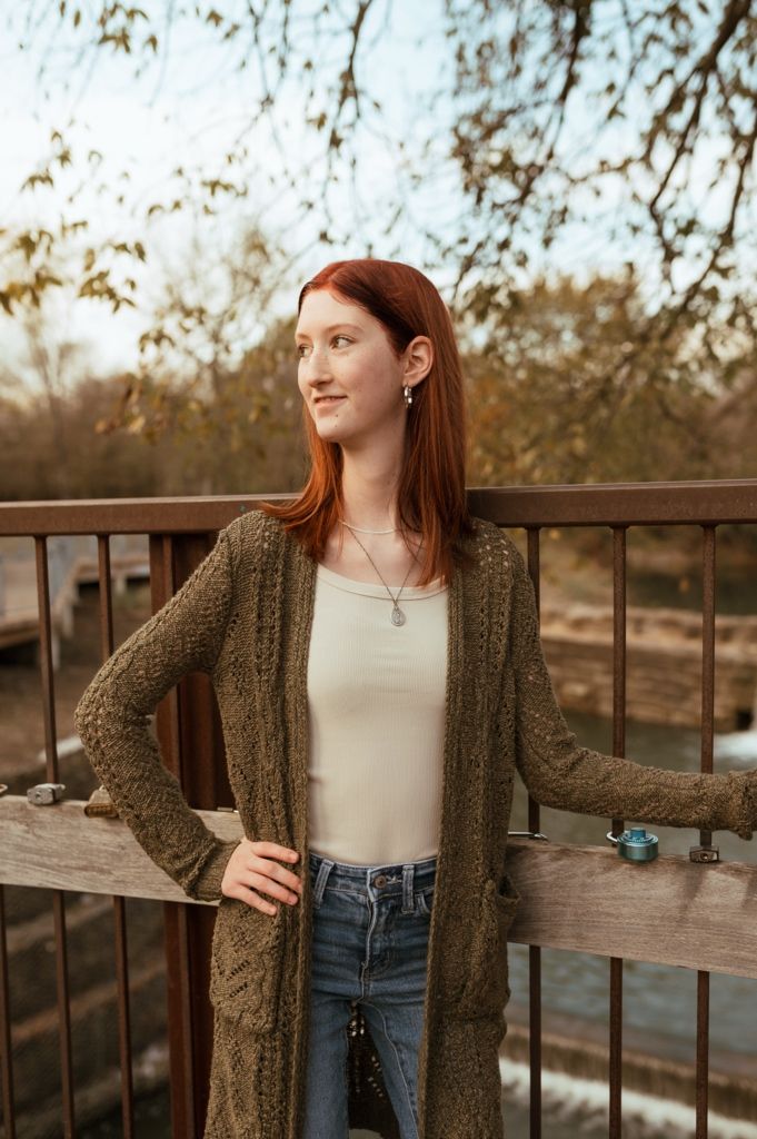 Person in olive cardigan and jeans poses against wooden railing during autumn sunset.