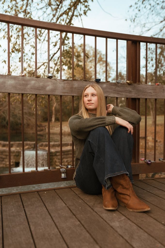 A person in boots and sweater sits contemplatively on wooden bridge deck.