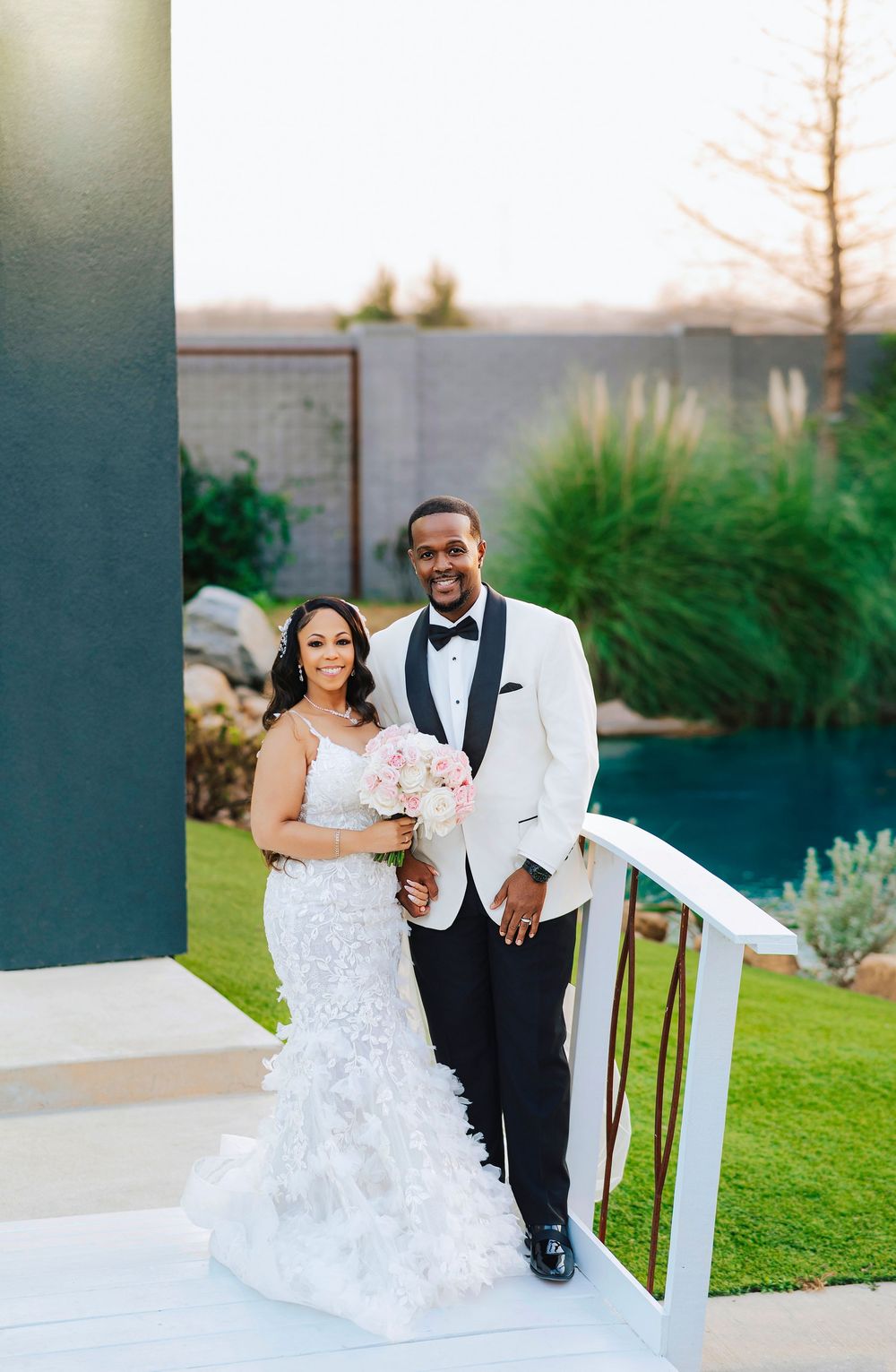 A couple in formal wedding attire pose together on a scenic outdoor terrace with greenery in the background.
