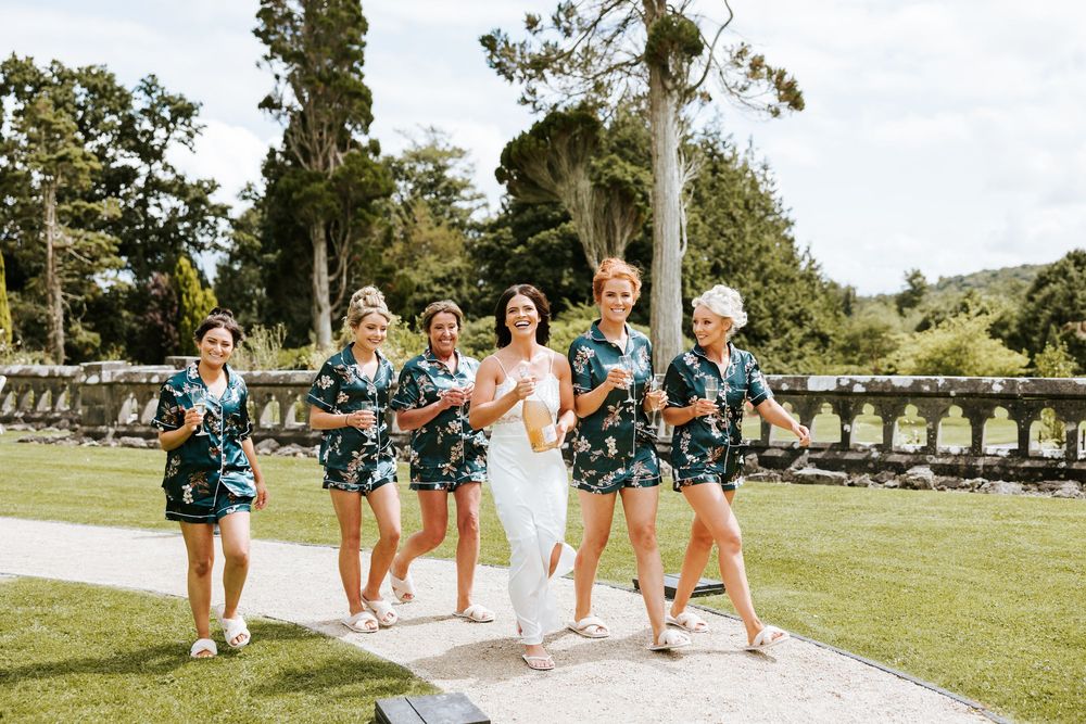 Bride in white dress walks with bridesmaids in matching floral robes on garden path at outdoor wedding. Markree Castle Sligo