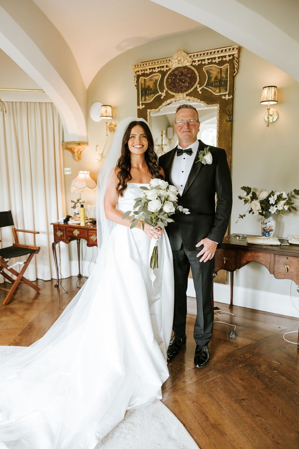 Bride in white gown holding bouquet stands with groom in tuxedo in elegant room with ornate mirror. Markree Castle Sligo