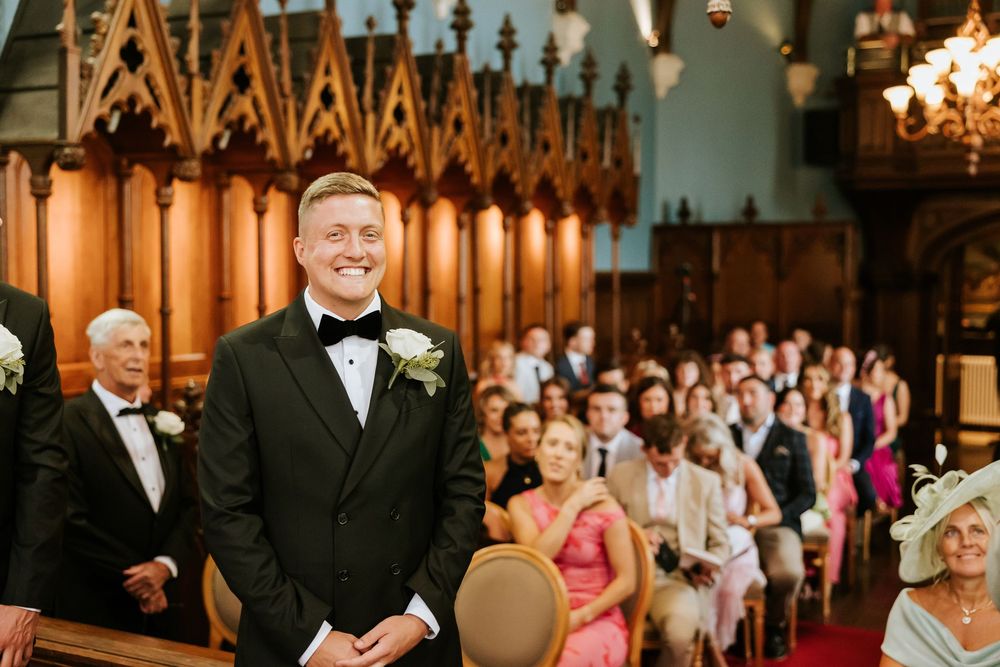 Groom in black tuxedo smiling at the altar inside an elegant church filled with wedding guests. Markree Castle Sligo
