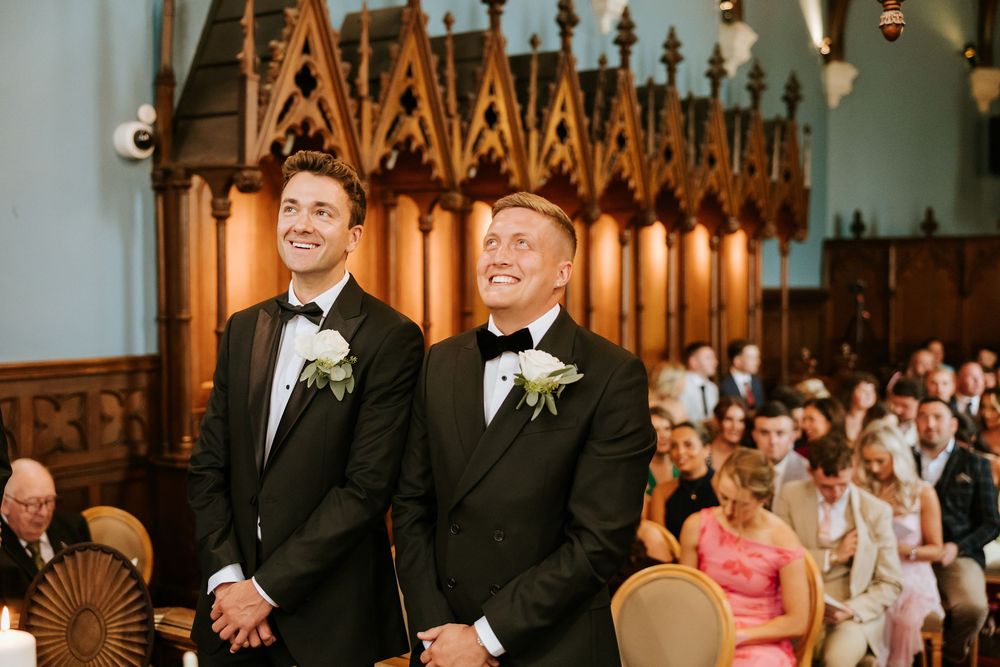 Two grooms in black tuxedos with white boutonnieres smile at the altar during their wedding ceremony in an elegant chapel. Markree Castle Sl