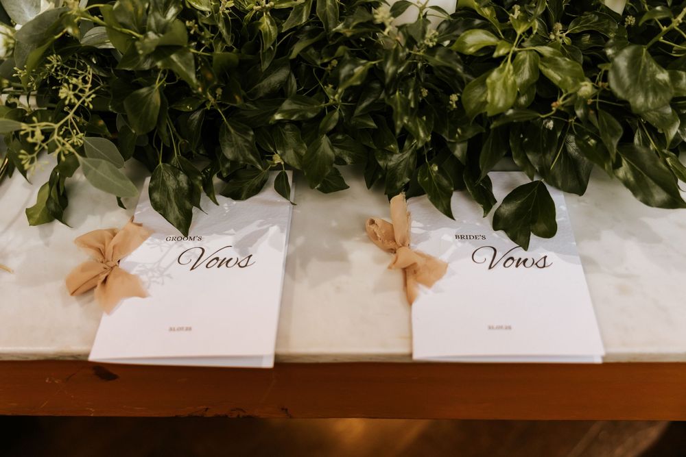 Two elegant white vow books with tan ribbon bows displayed on a wooden table with lush green floral backdrop. Markree Castle Sligo