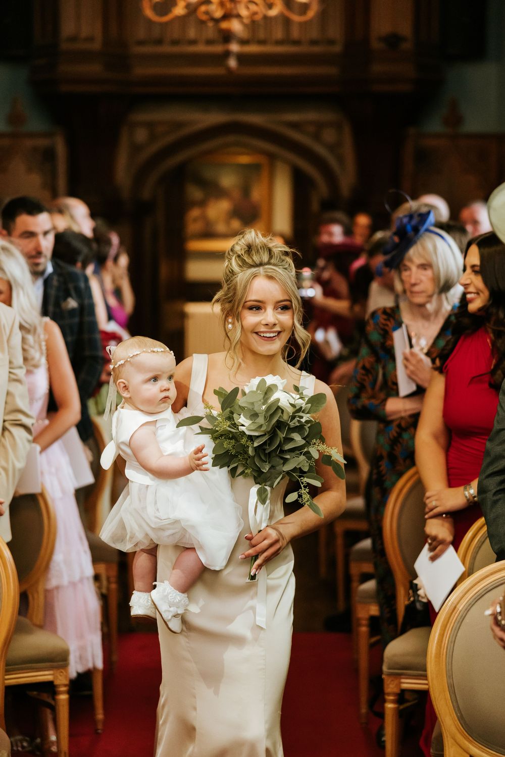 Bridesmaid walks down the aisle carrying a baby and white bouquet in an elegant wedding ceremony. Markree Castle Sligo