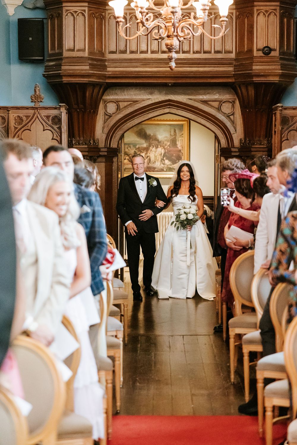 Bride and groom walk down the aisle at an elegant wood-paneled wedding ceremony venue with guests seated beside them. Markree Castle Sligo